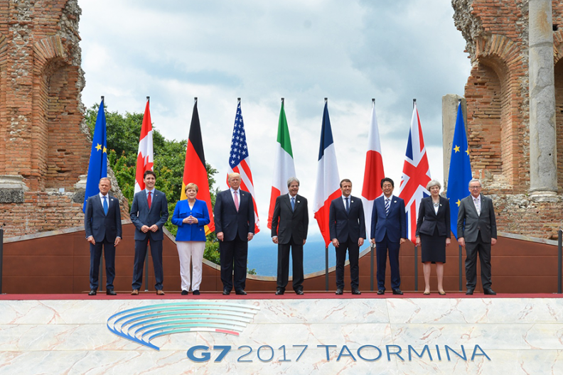 Szczyt państw G7 w Taorminie (Włochy, 2017 r.). Od lewej: Donald Tusk, Justin Trudeau, Angela Merkel, Donald Trump, Paolo Gentiloni, Emmanuel Macron, Shinzō Abe, Theresa May i Jean-Claude Juncker na tle flag Unii i flag państw G7. / Źródło: Wikimedia